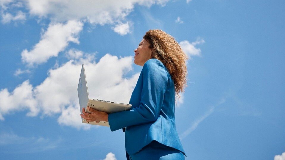 Mujer de pie con portátil abierto bajo un cielo azul nublado.